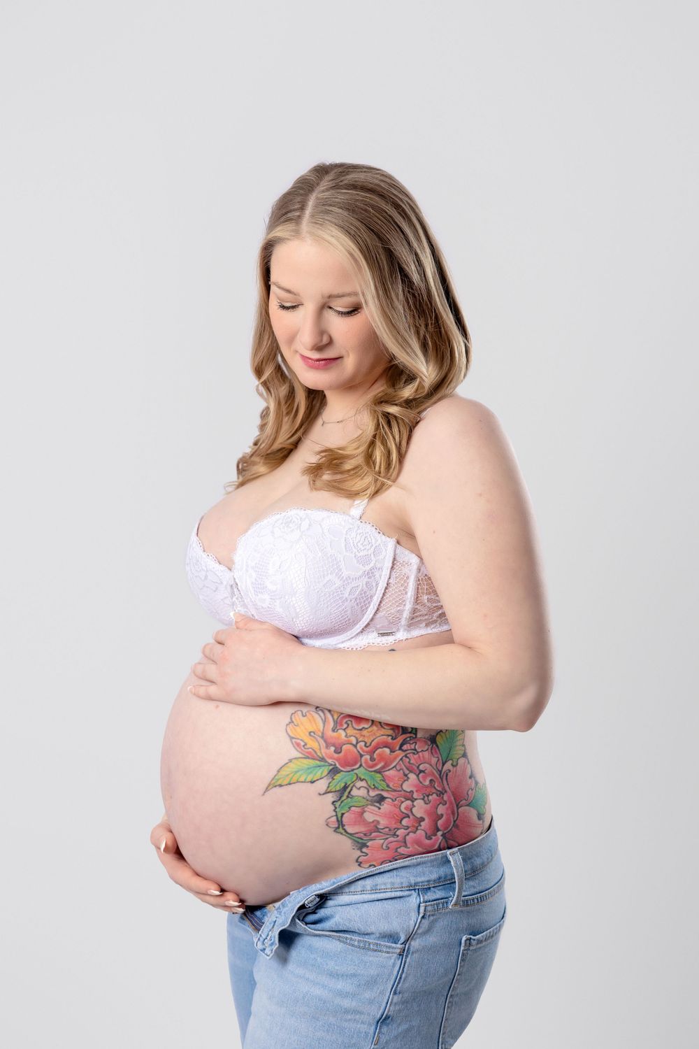 Side profile of an expecting parent in jeans and white top, showing a colorful floral tattoo on their belly.