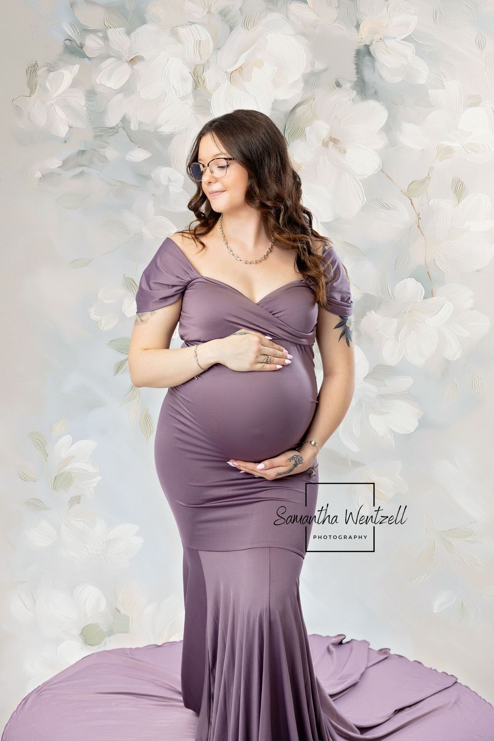 A maternity photo featuring a purple off-shoulder gown and floral backdrop in soft, ethereal lighting.