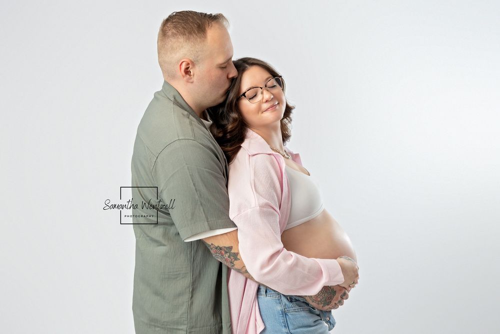 Expectant parents embrace against white background, wearing green shirt and pink cardigan with jeans.