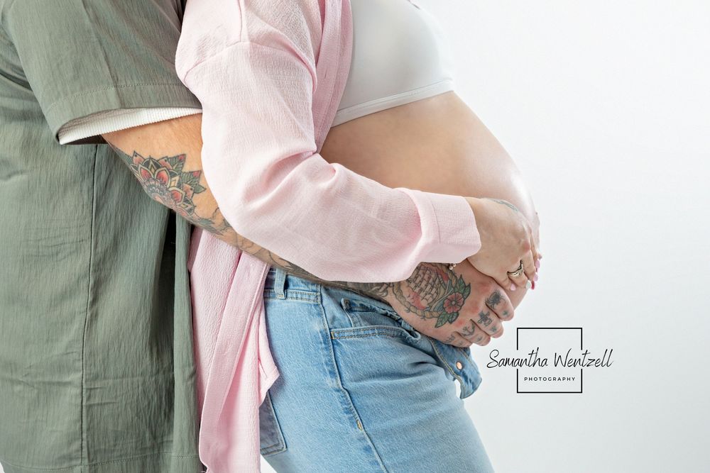 Side view close up of pregnant belly being cradled by tattooed hands in intimate maternity pose.