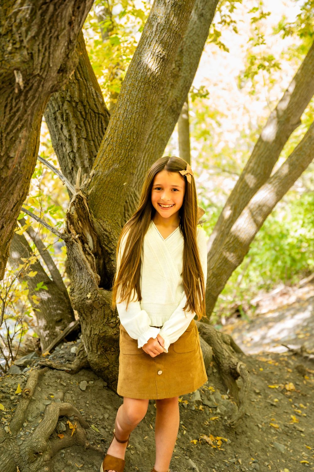 Young child poses playfully near a large tree trunk in natural sunlight.