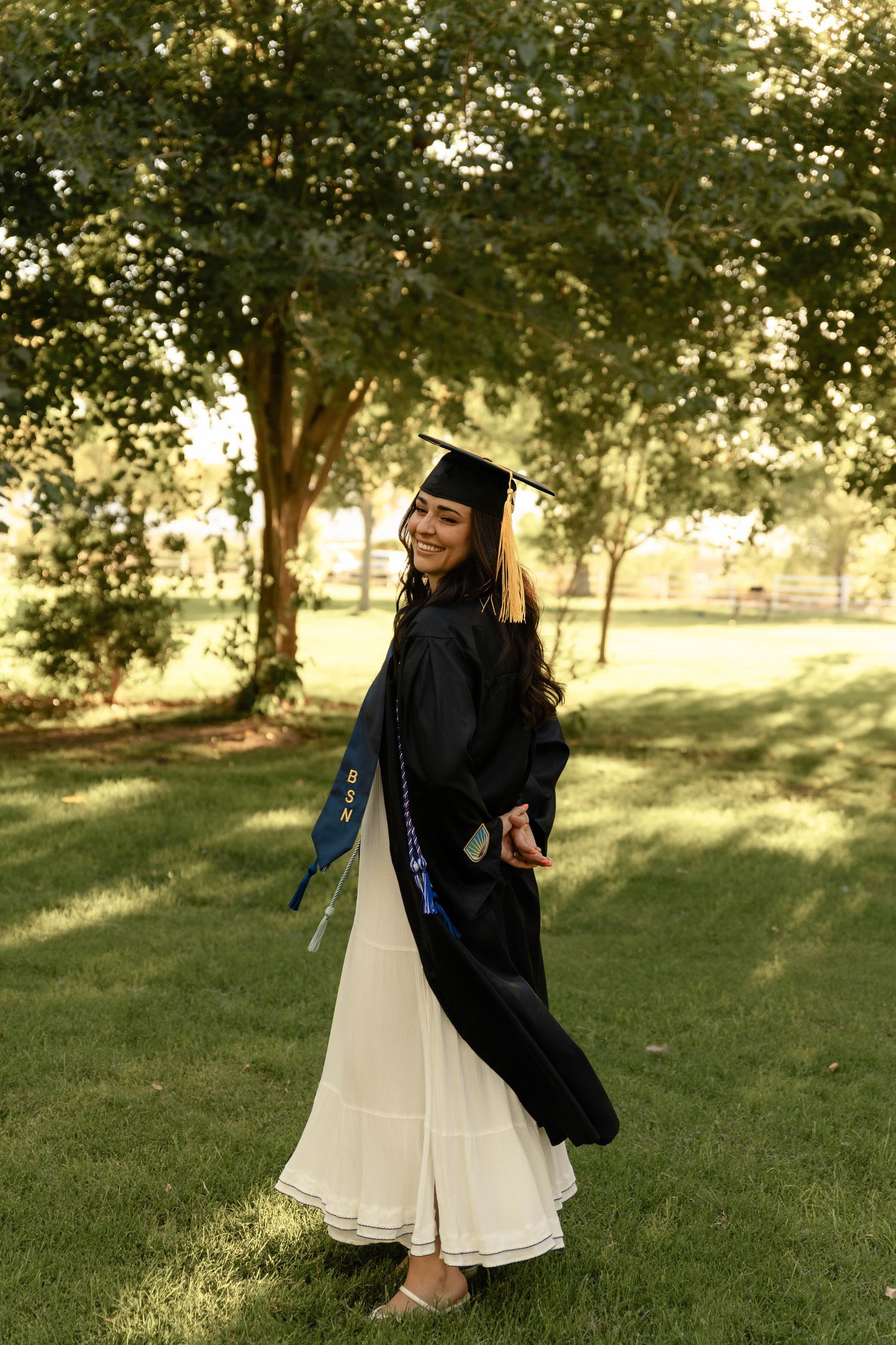 Smiling woman in black graduation cap and gown posing outdoors among green trees.
