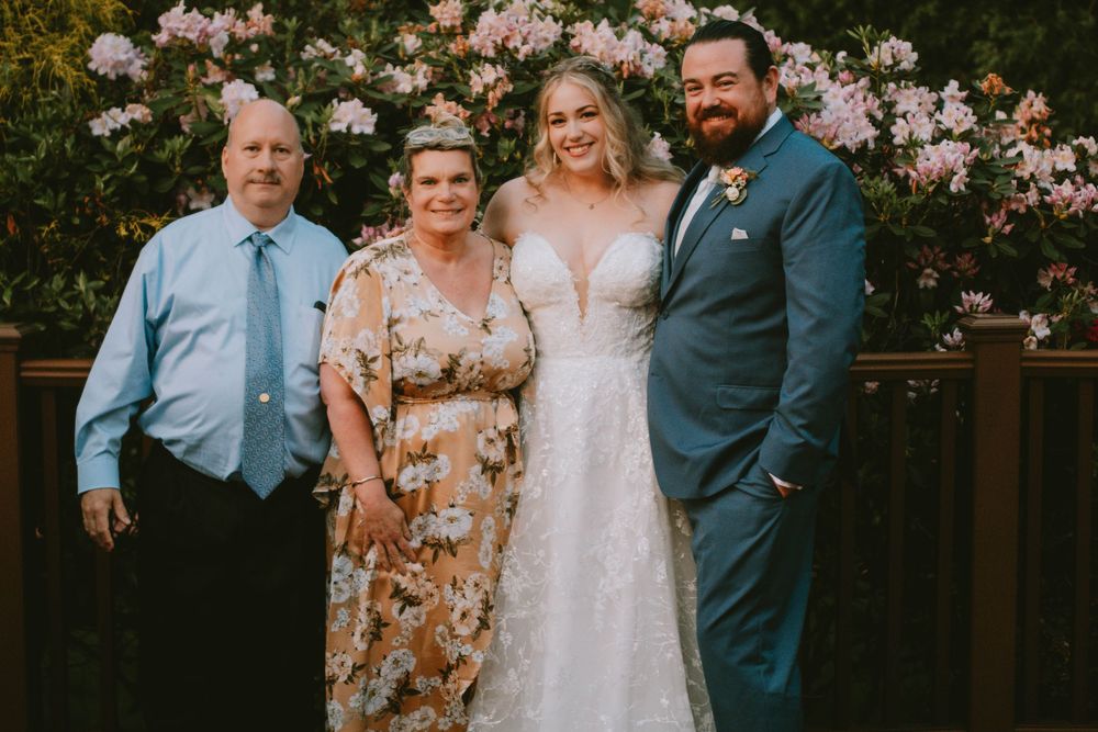 Wedding party group photo with floral backdrop of pink roses in a garden setting.
