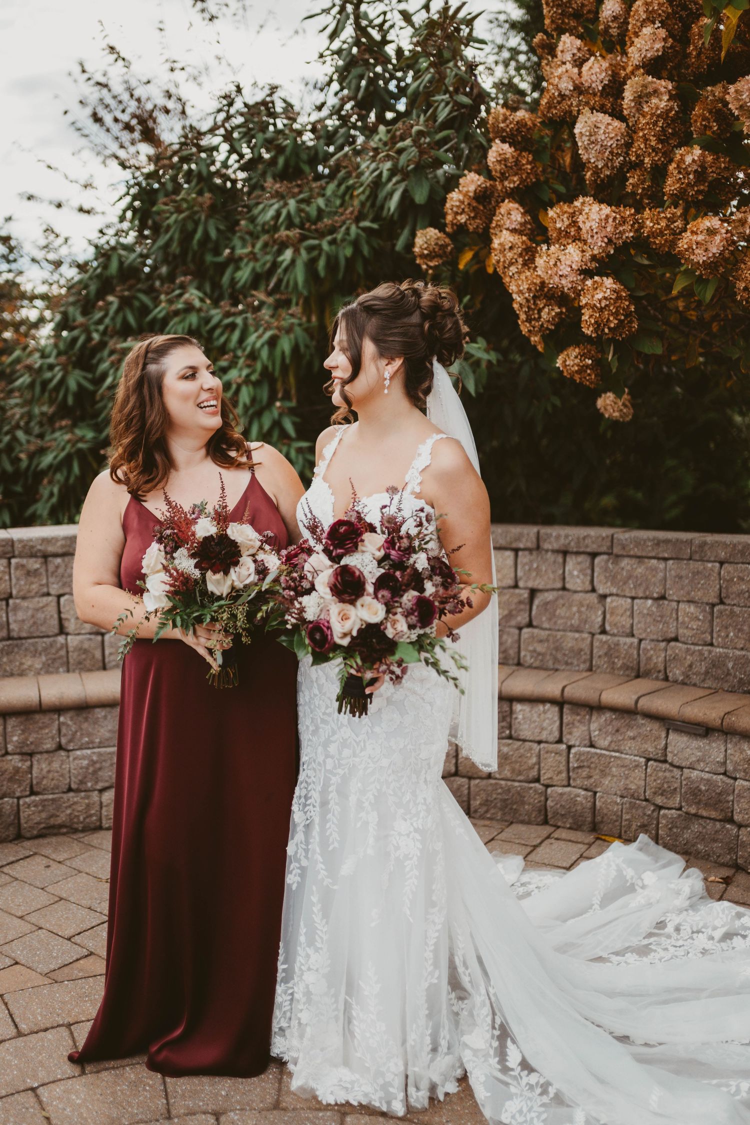 Wedding party members pose with burgundy bouquets against a stone wall and autumn foliage.