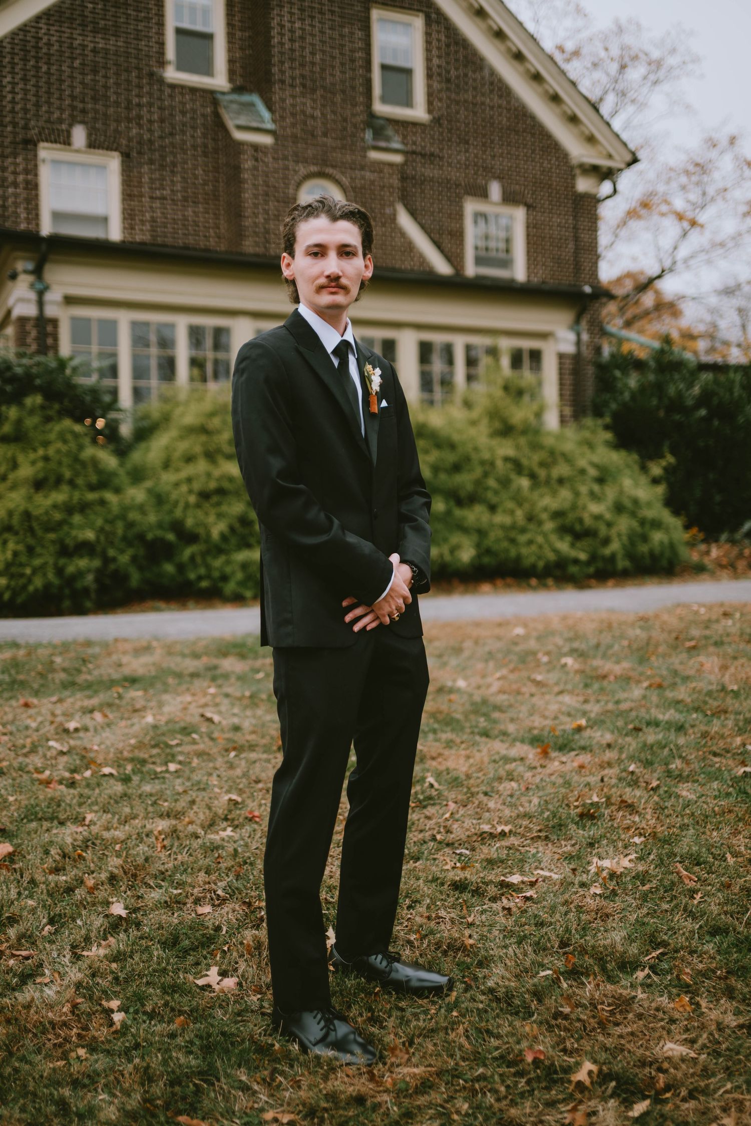A groom in a dark suit stands on a lawn in front of a brick house.