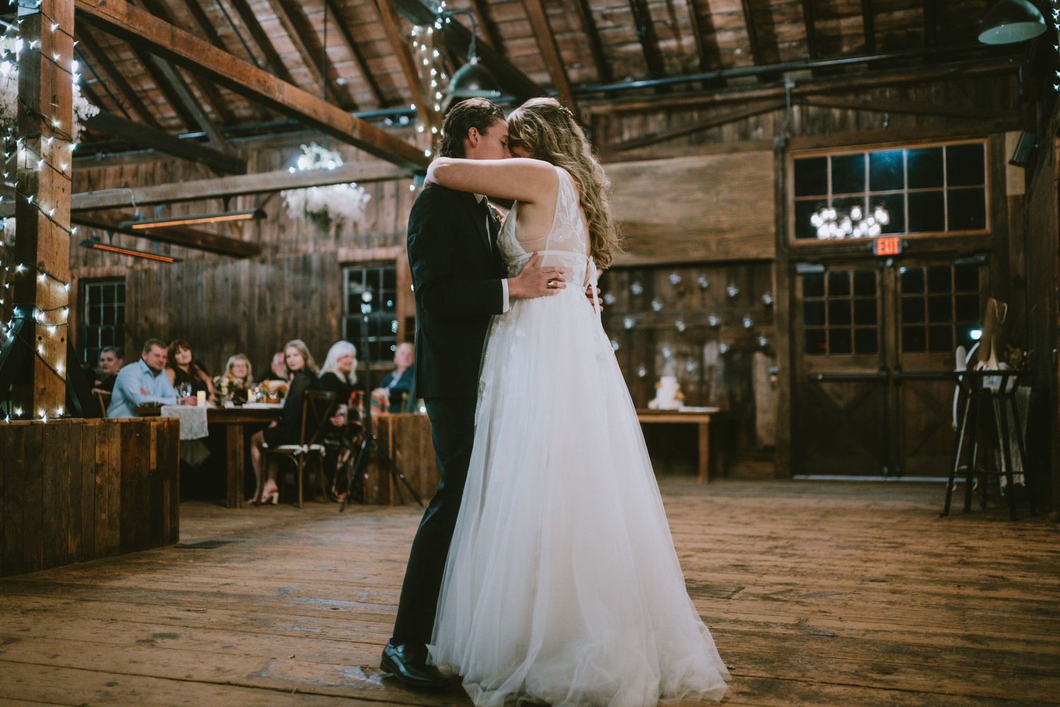 A couple shares their first dance in a rustic barn venue with wooden beams and string lights.