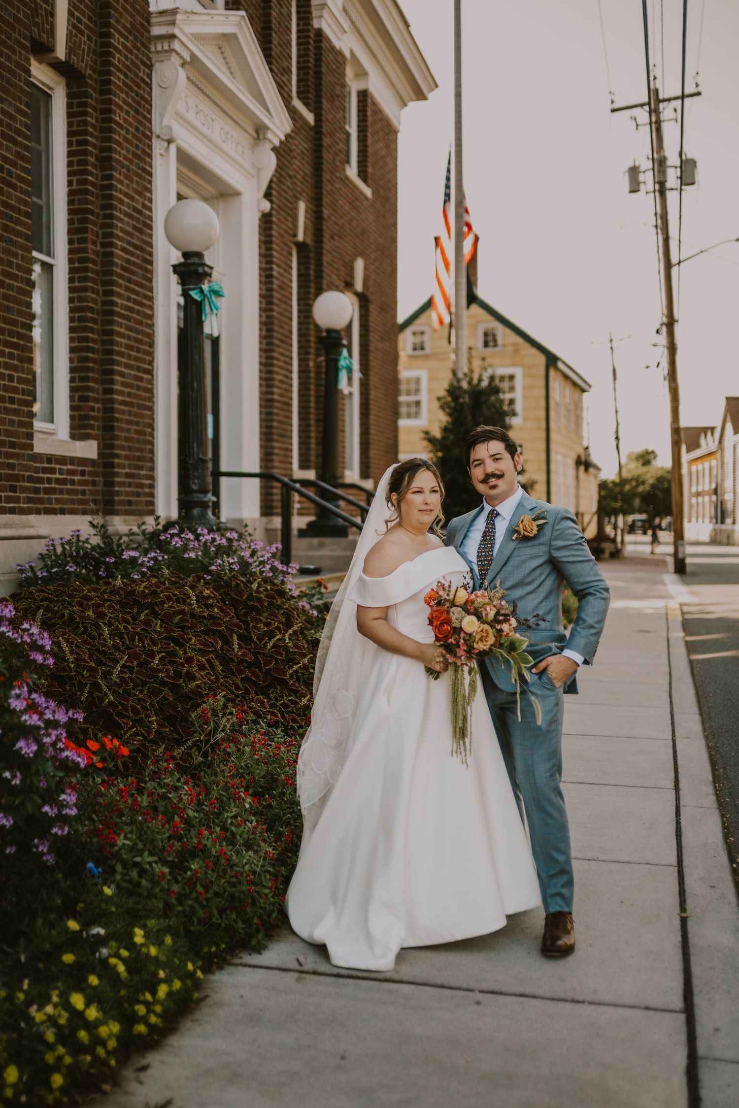 A couple poses for wedding photos on a sidewalk with brick buildings and colorful flowers.