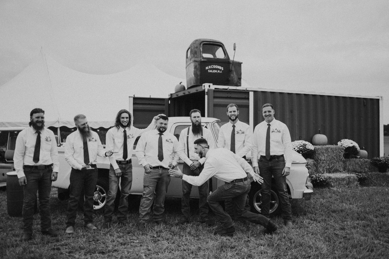 Wedding party poses in black and white photo with vintage vehicles in background.
