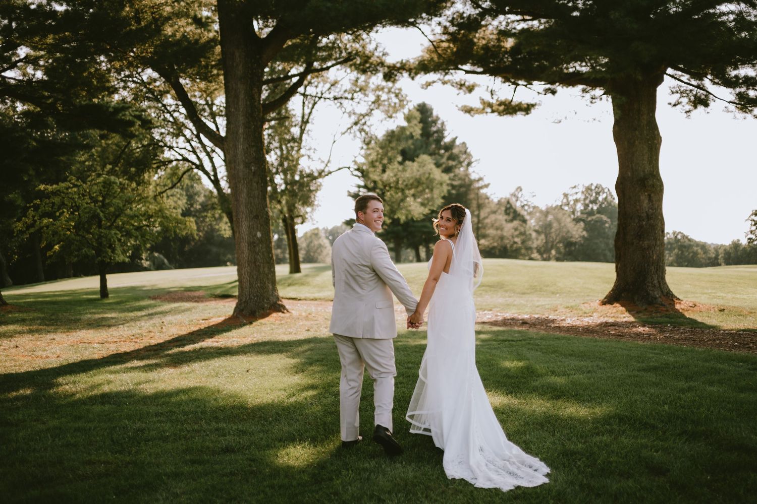 A bride and groom hold hands on a lush green lawn surrounded by tall trees during their outdoor wedding ceremony.