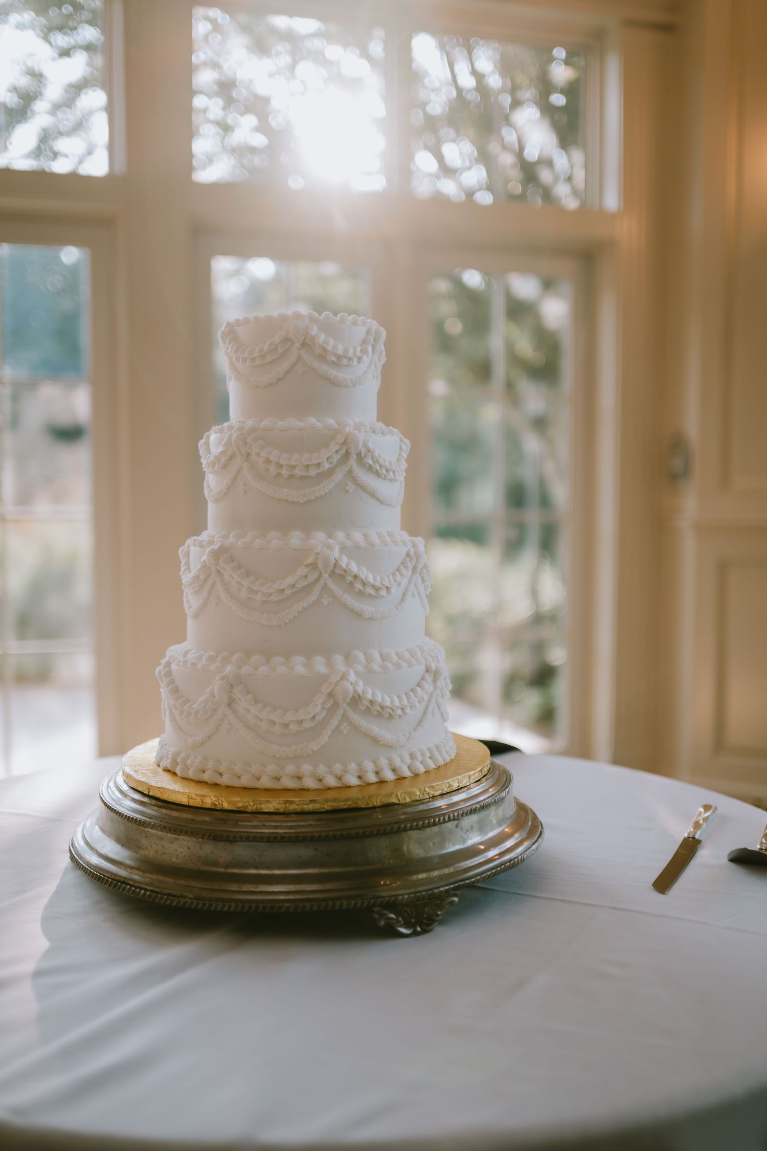 Elegant four-tier white wedding cake with pearl and swag piping on a gold stand by a sunlit window.