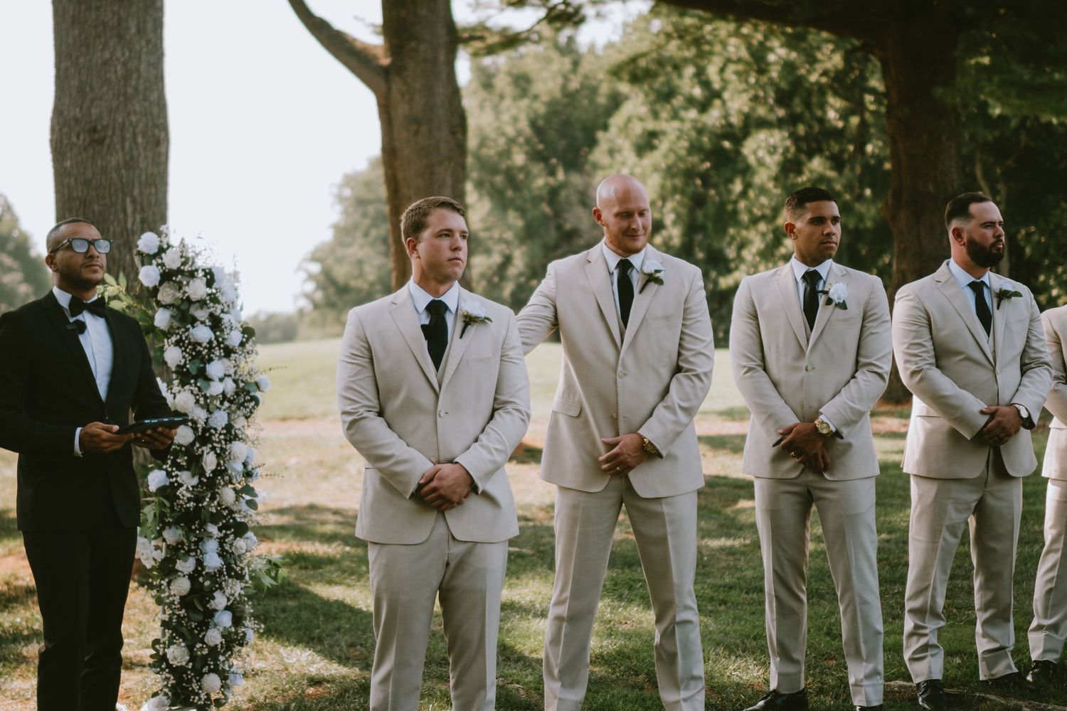 Groomsmen in tan suits stand outdoors at a wedding ceremony beside a floral arch with white flowers.