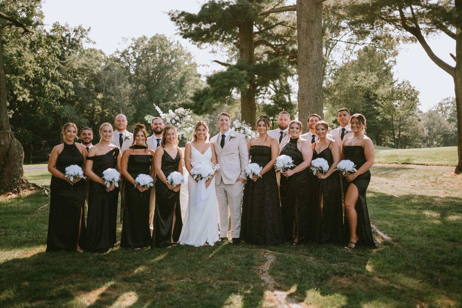 Wedding party posing outdoors, bridesmaids in black gowns holding white bouquets flanking bride and groom in white and tan.