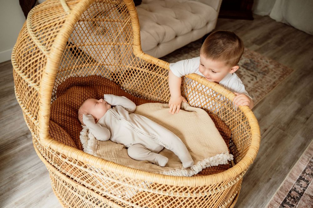 Newborn baby sleeping in a wicker bassinet while an older toddler sibling watches over lovingly.