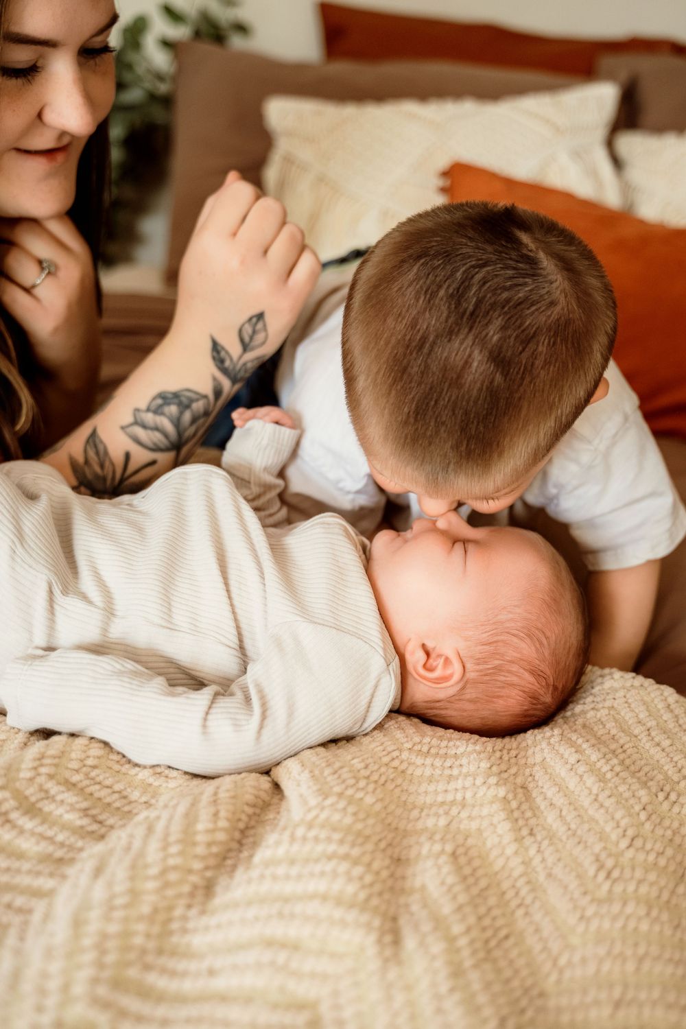 A tattooed mother smiles as her young son gently kisses their newborn baby lying on a cozy bed.
