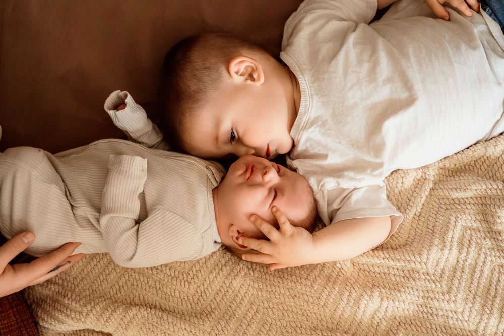A toddler tenderly cuddles a sleeping newborn baby on a cozy blanket, capturing a sweet sibling bonding moment.