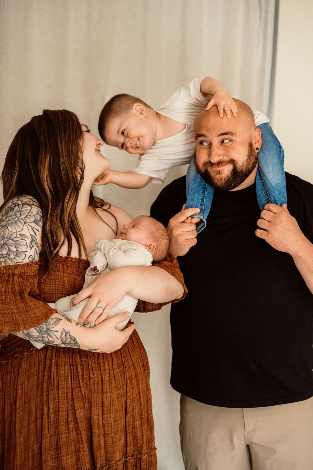 A tattooed mother holds a newborn while a toddler sits on the smiling bald father's shoulders in a warm family portrait.