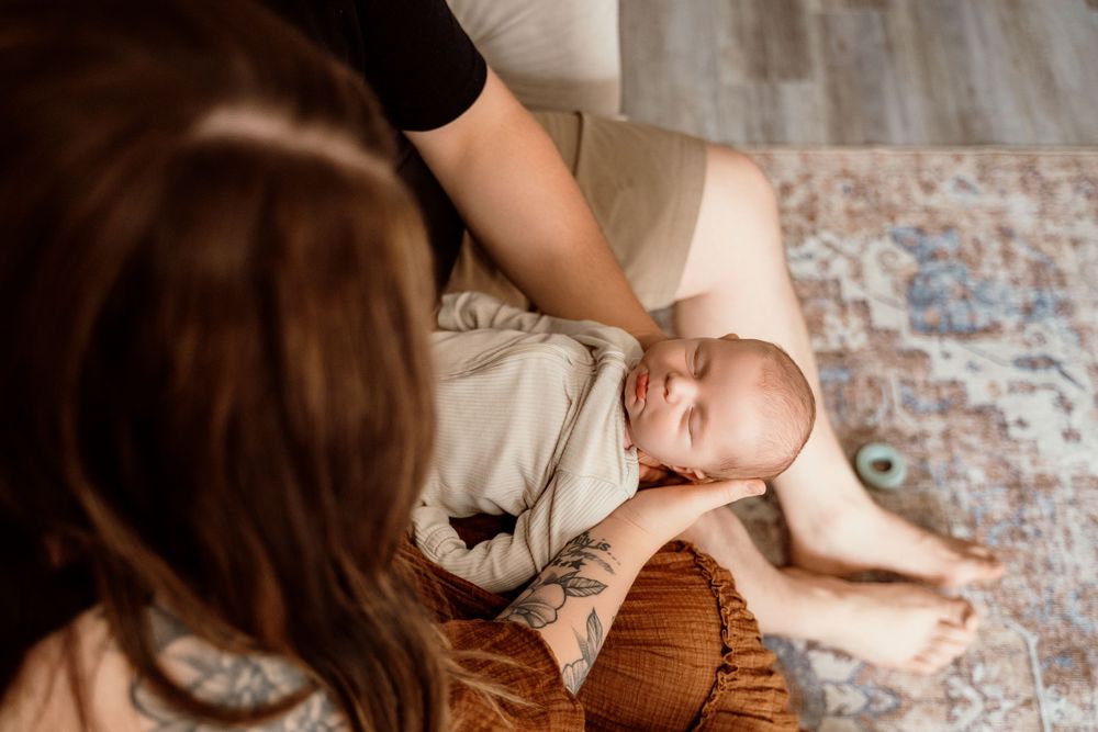 A mother sits on a rug cradling her newborn baby wrapped in a white swaddle blanket during an indoor newborn photo session.