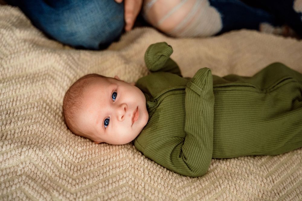 Baby wearing green long sleeve onesie lies on beige textured blanket looking up at camera.