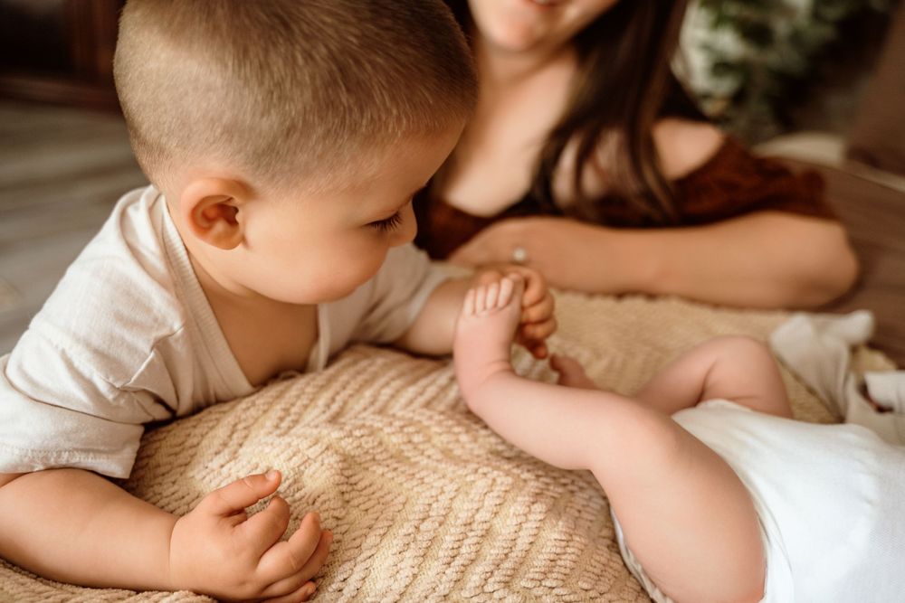 A toddler in a white onesie curiously looks at a newborn baby lying on a woven blanket with a mother nearby.