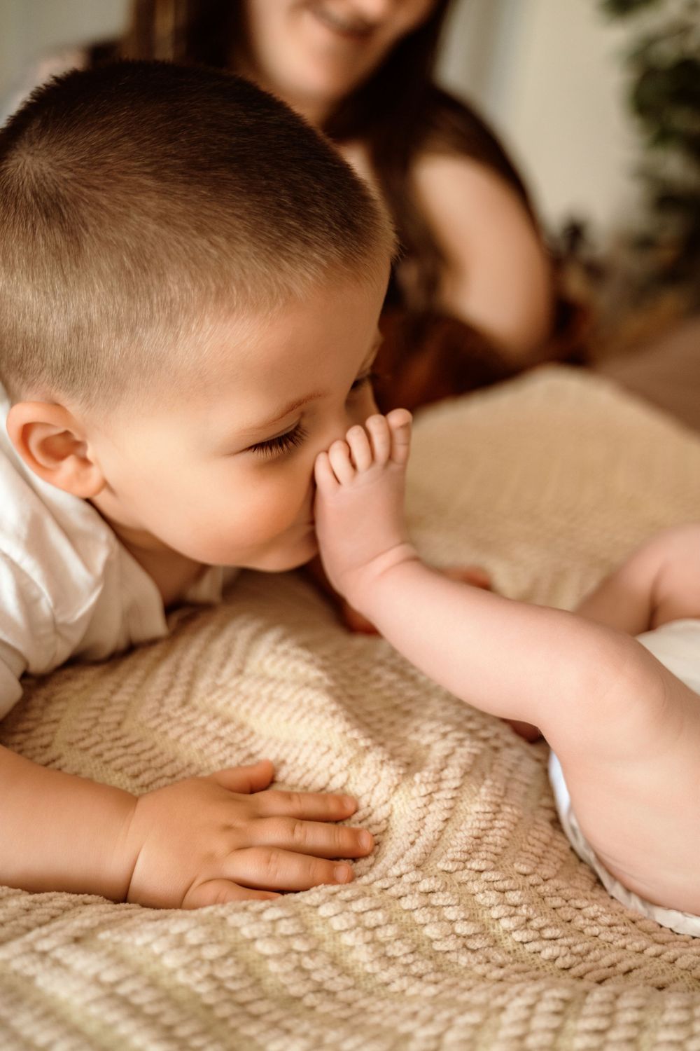 A toddler boy gently kisses a newborn baby's foot on a beige knit blanket while a smiling mother watches in the background.