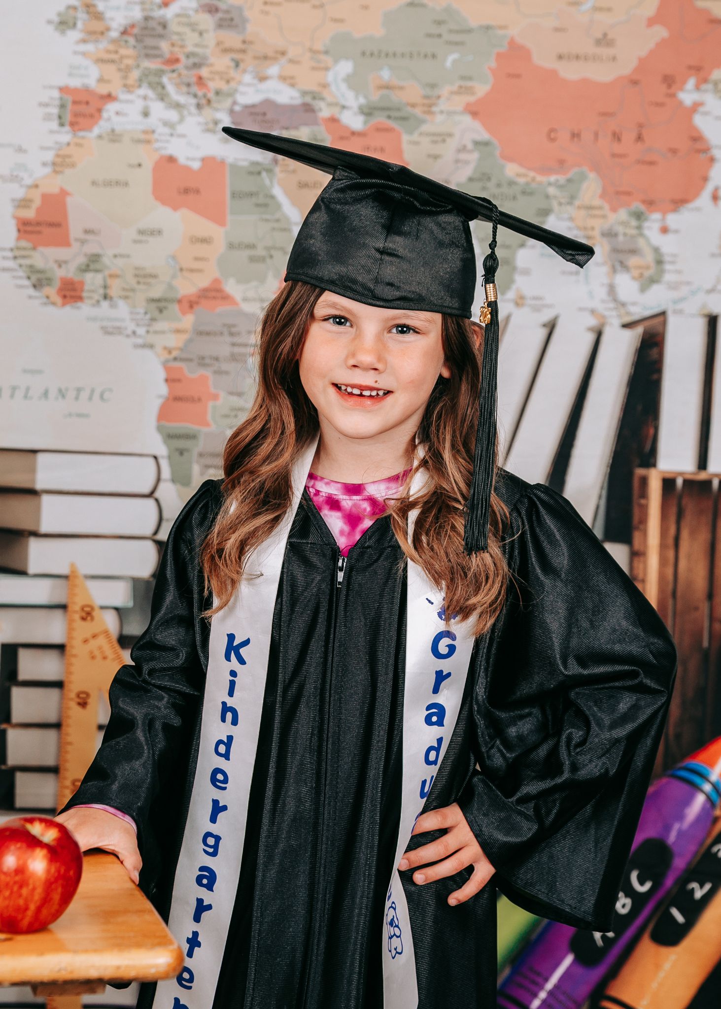 A young girl proudly wears a black graduation cap and gown with a Kindergarten Graduate sash indoors.