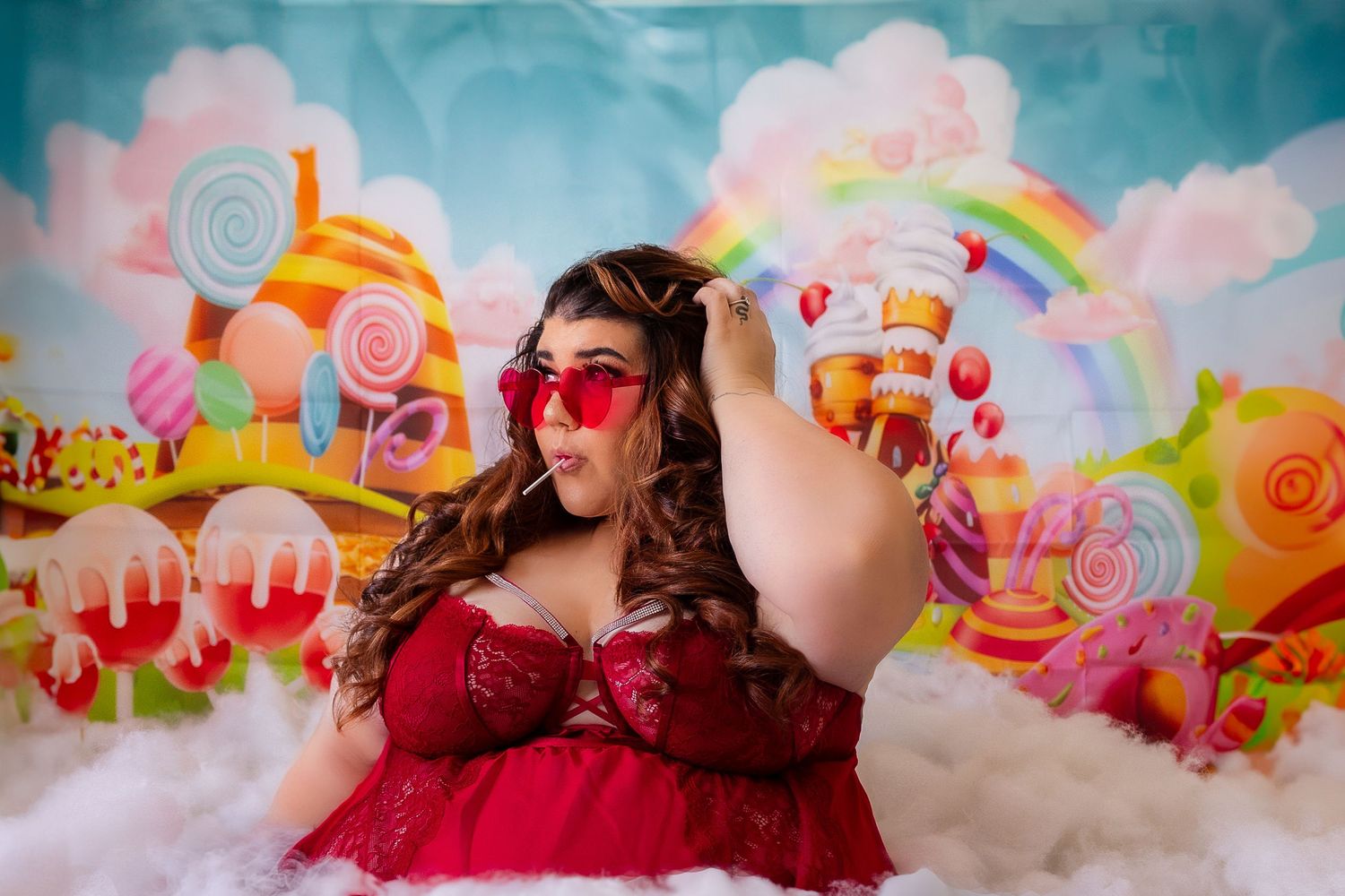 Fantasy candy themed portrait with lollipops, clouds, and rainbow background featuring model in red dress and sunglasses.