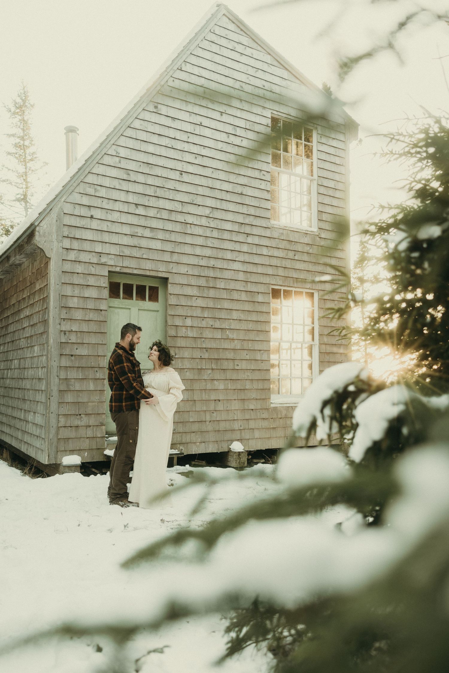 A couple stands together outside a snow-covered cabin in winter sunlight.