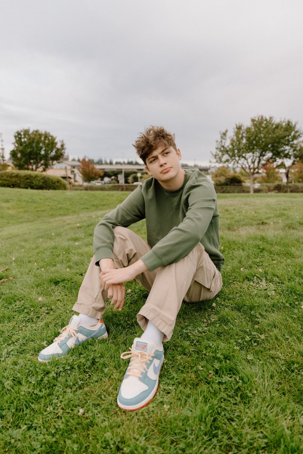 Casual portrait of someone in a green hoodie sitting on grass wearing colorful sneakers.