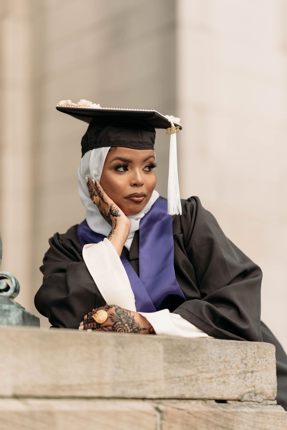 Graduate in academic regalia and hijab poses thoughtfully on stone steps.