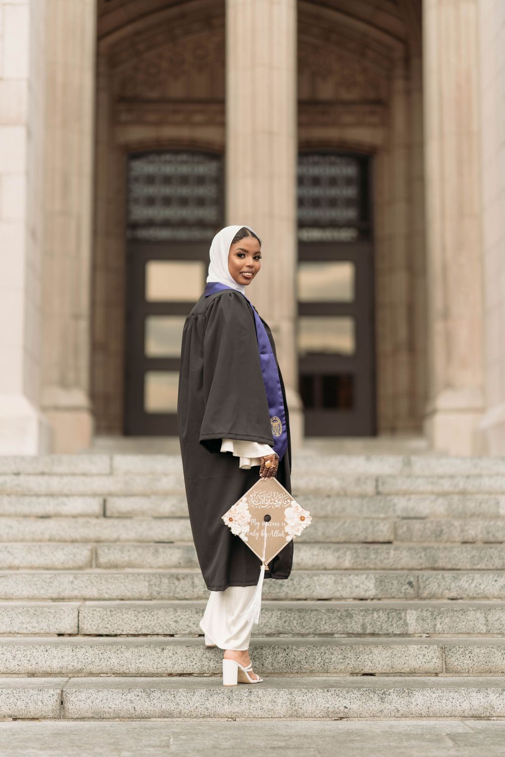 Graduate in traditional hijab and academic regalia poses on stone steps of building.