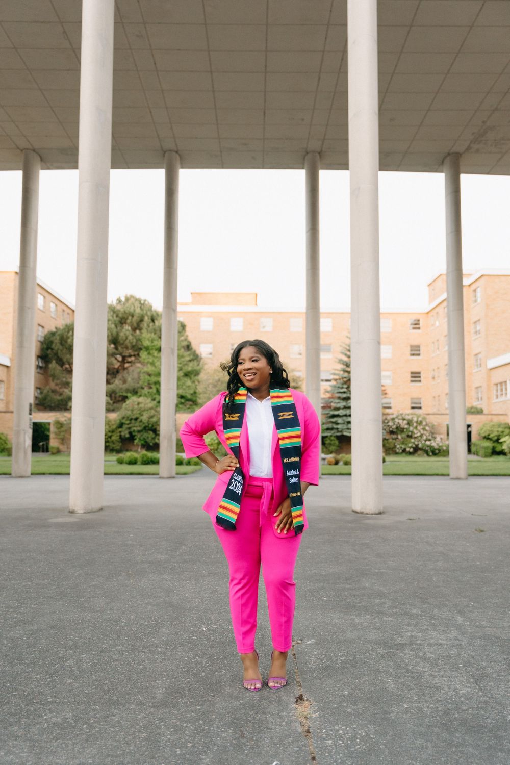 Graduate in bright pink suit poses confidently under white columned pavilion.