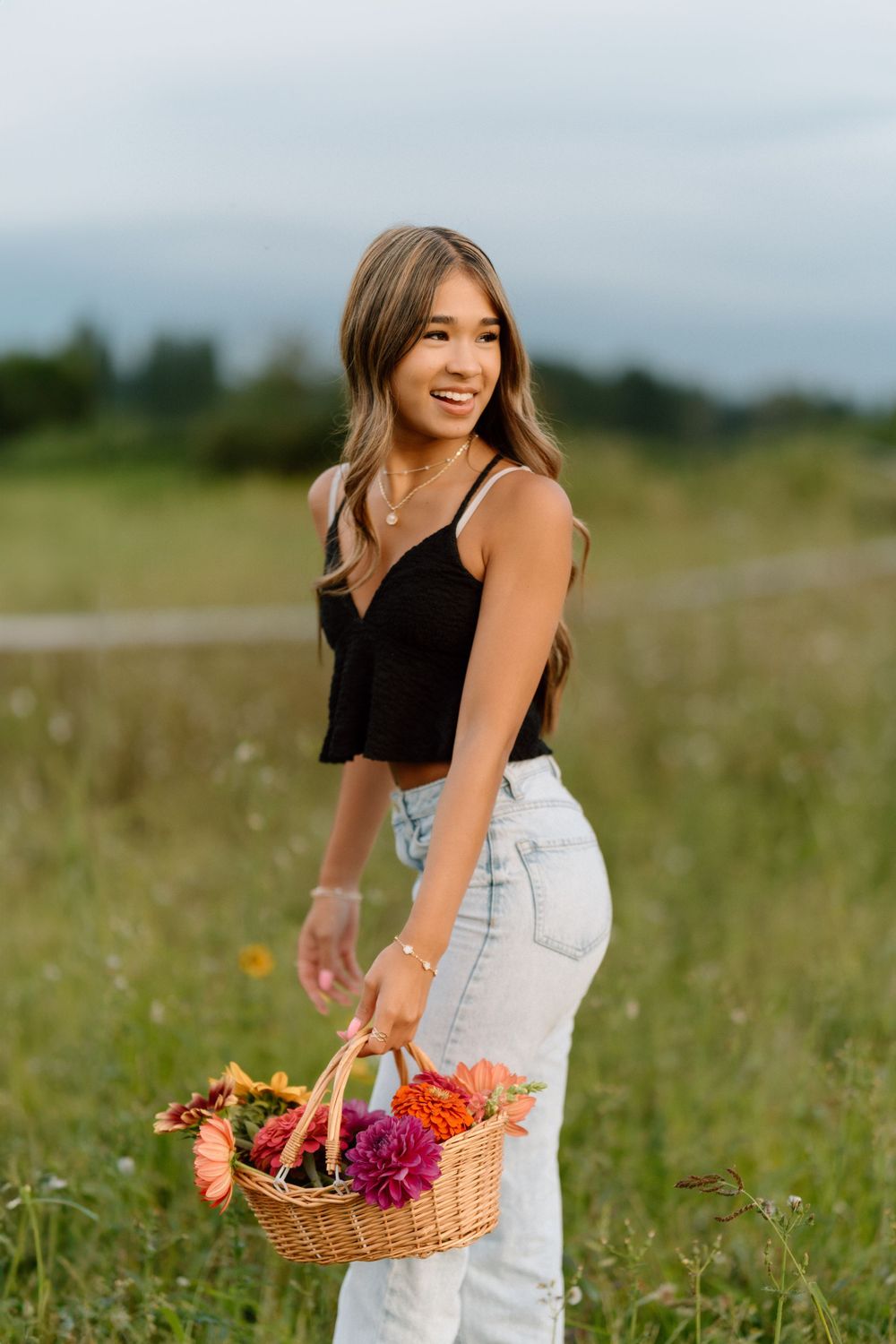 A person in casual attire holds a basket of fresh flowers in a summer field.
