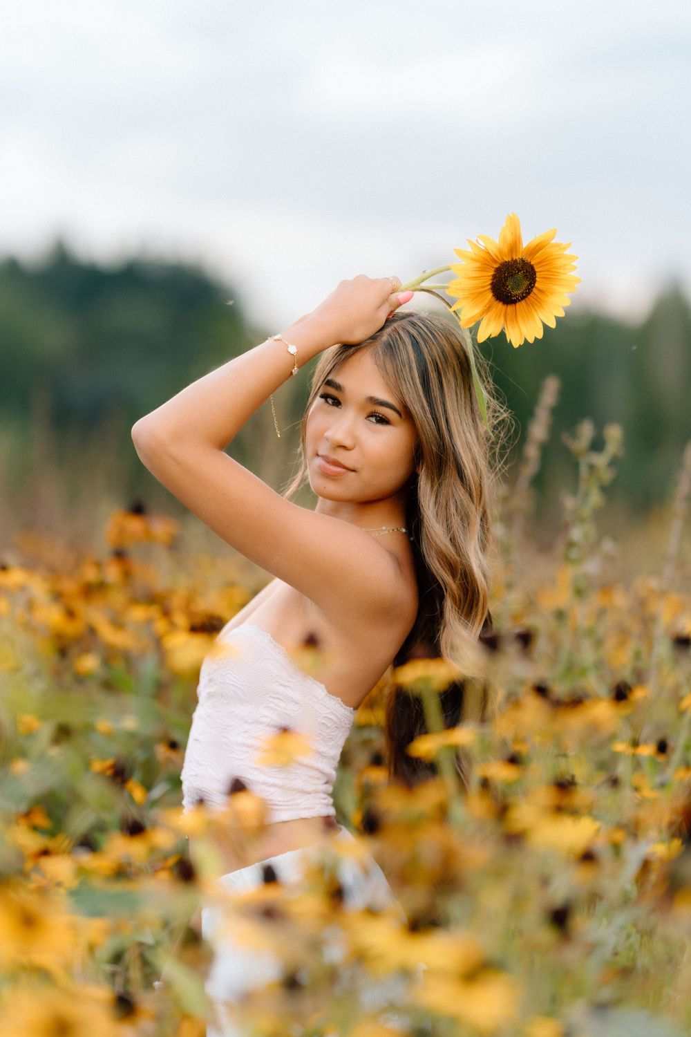 A person poses with a sunflower in a golden sunflower field during sunset.