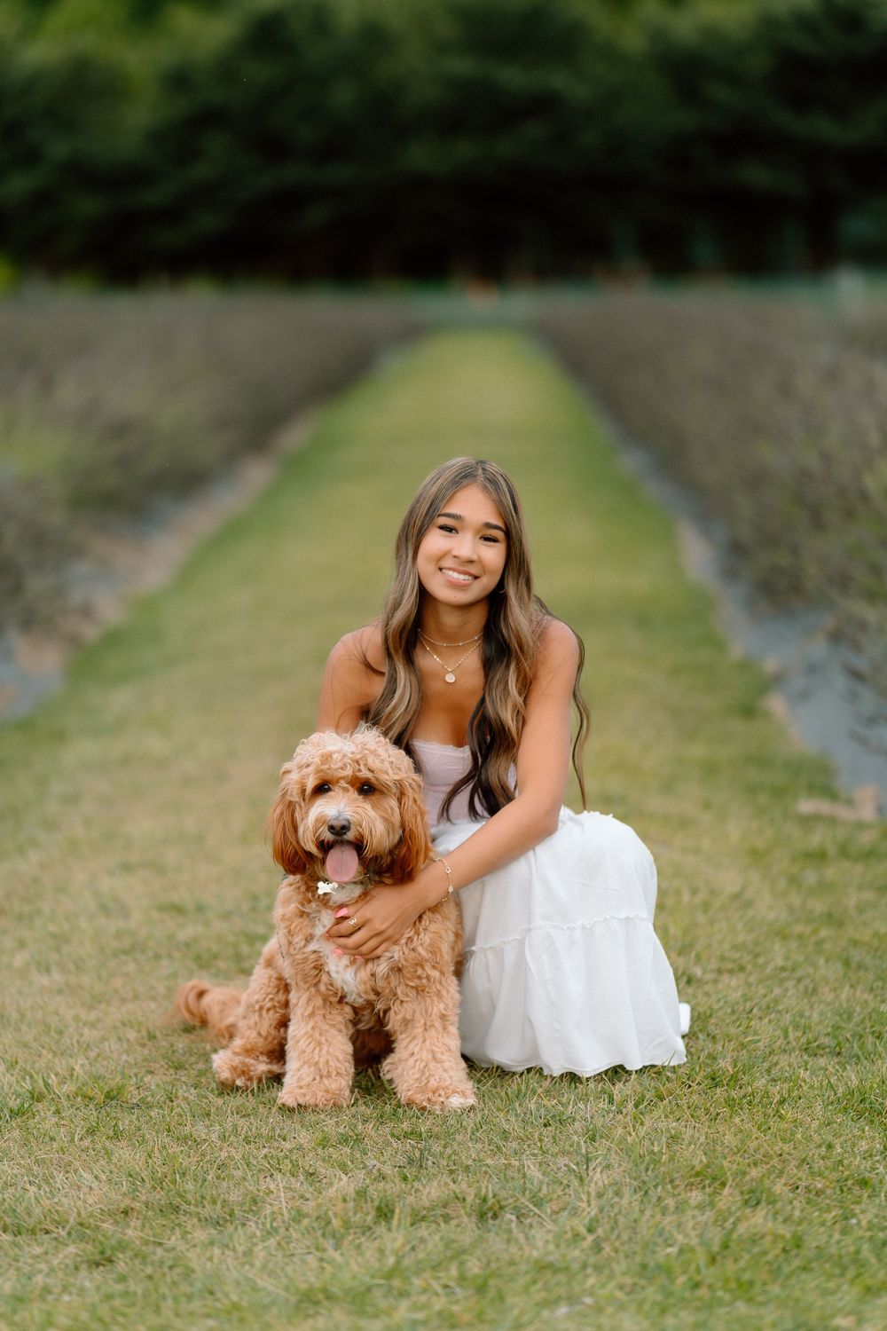 A person in a white dress poses with a friendly goldendoodle dog on a grassy path.
