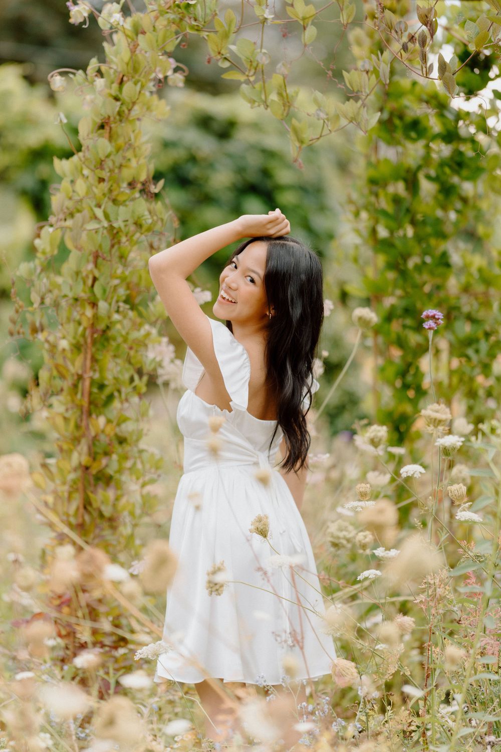 A person in a white summer dress poses gracefully among blooming flowers and vines.