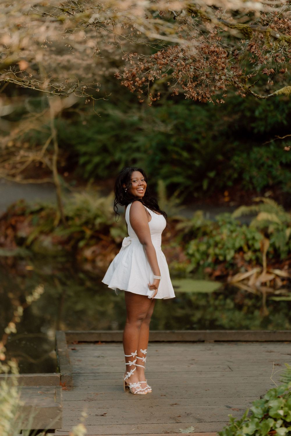 Someone in a white sundress poses on a wooden deck surrounded by lush garden scenery.