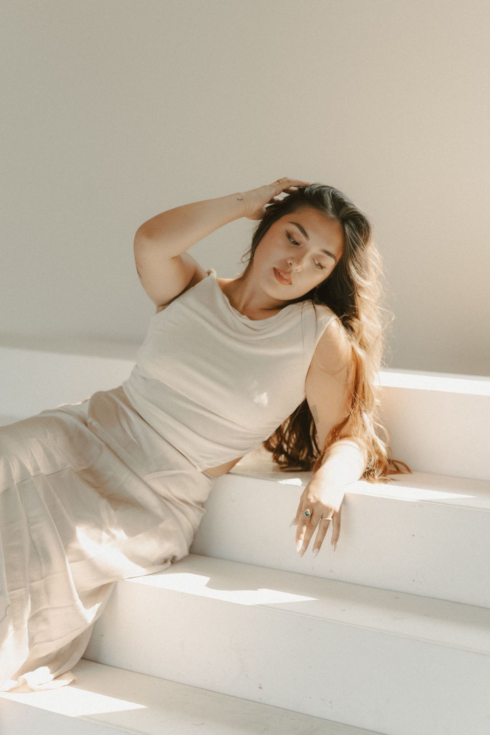 A woman in white lounges on sunlit stairs in a minimalist setting.