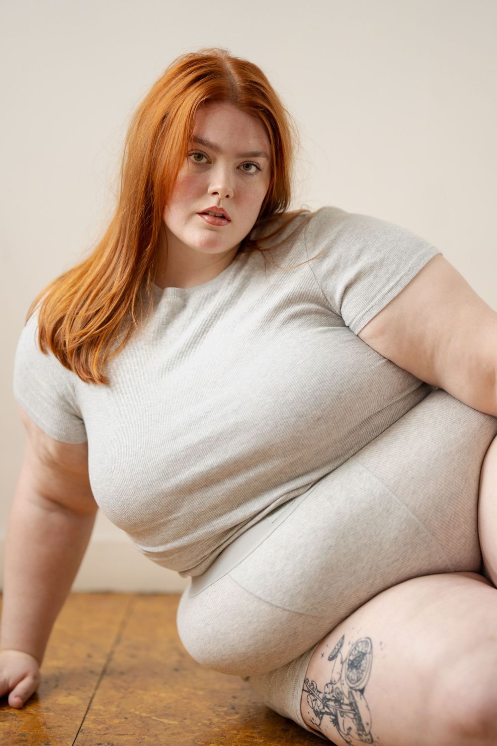 Model wearing light grey loungewear set sitting on wooden floor showing body confidence.