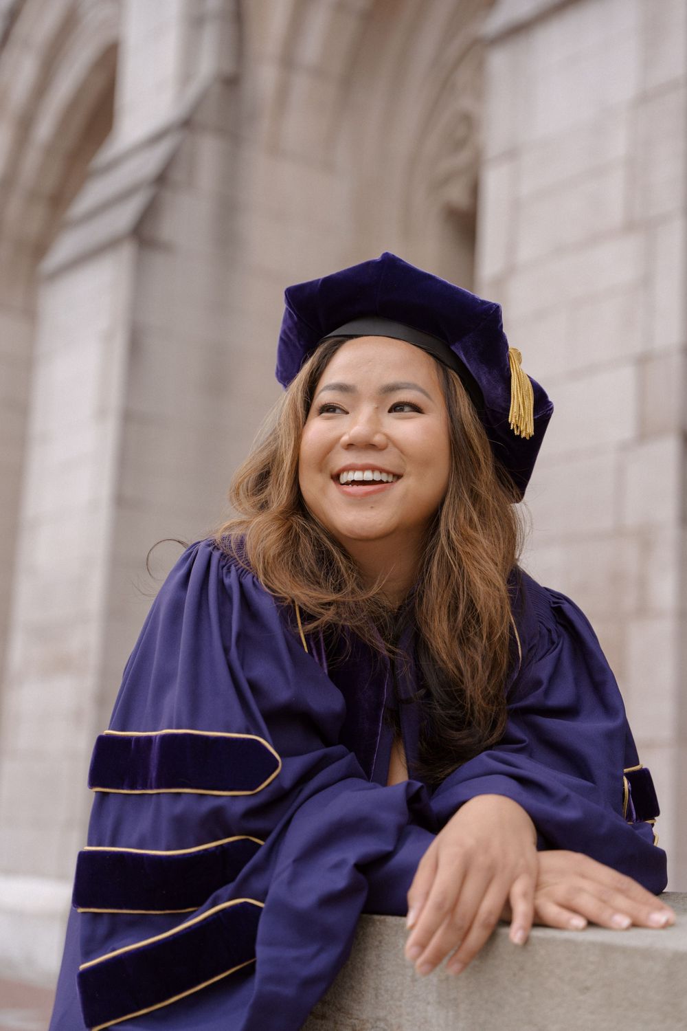 Graduate in purple doctoral regalia and cap smiles while leaning on stone ledge.
