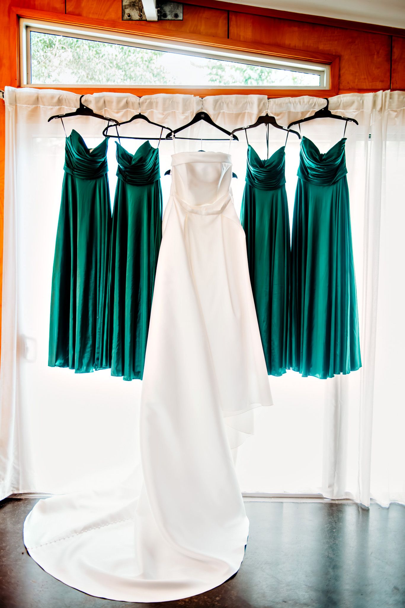 A white wedding gown hangs alongside four emerald green bridesmaid dresses by a window.