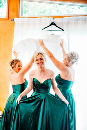 Bridesmaids in emerald green dresses dance and celebrate together in a sunlit room.