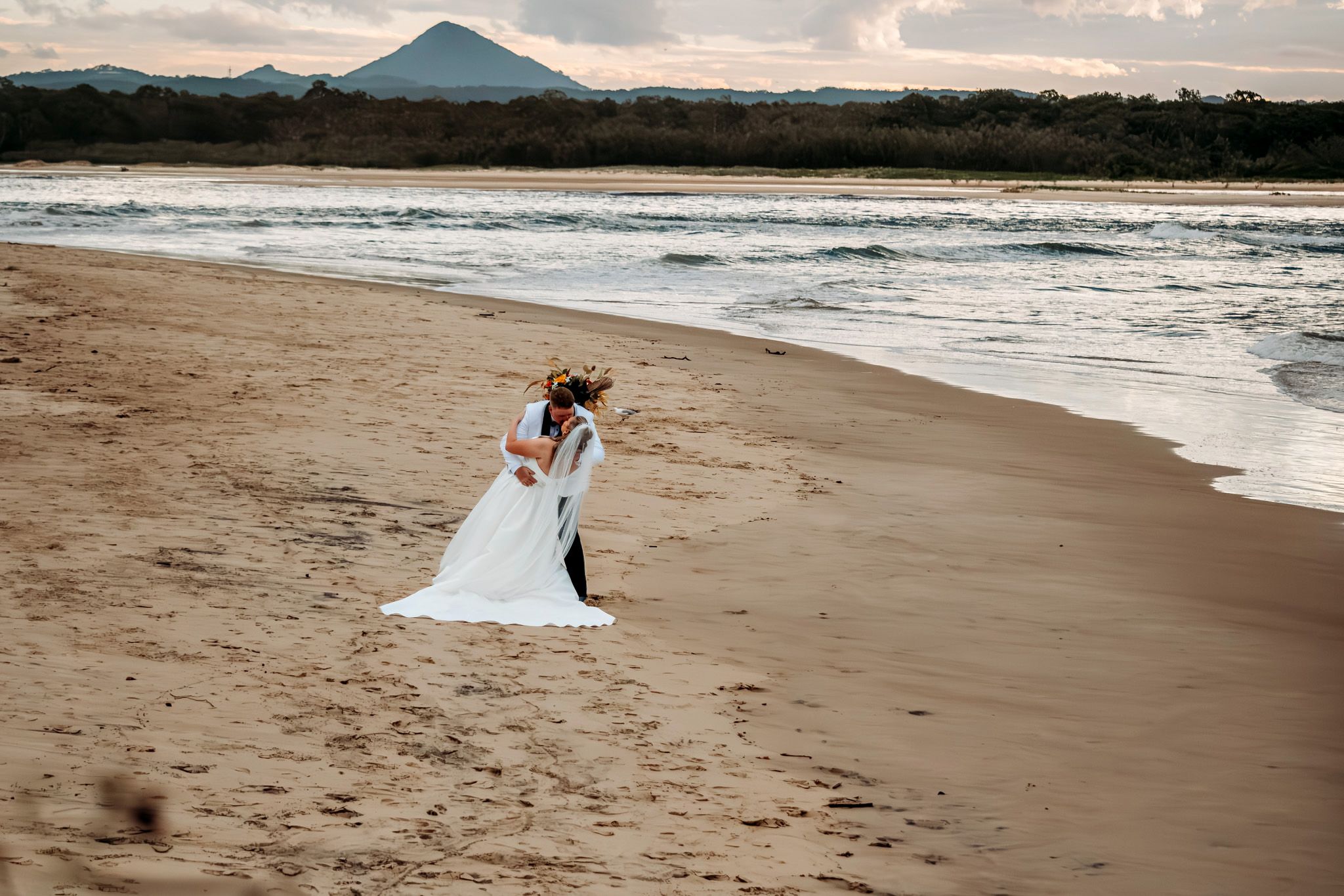 Newlyweds walking along a beach with a mountain silhouette in the background.