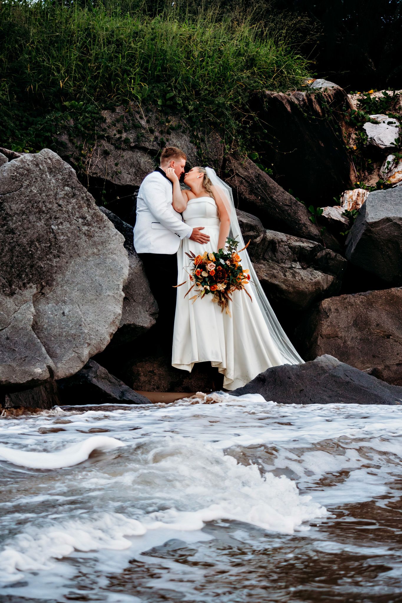 A wedding couple embraces on rocky coastal formations as waves crash below.