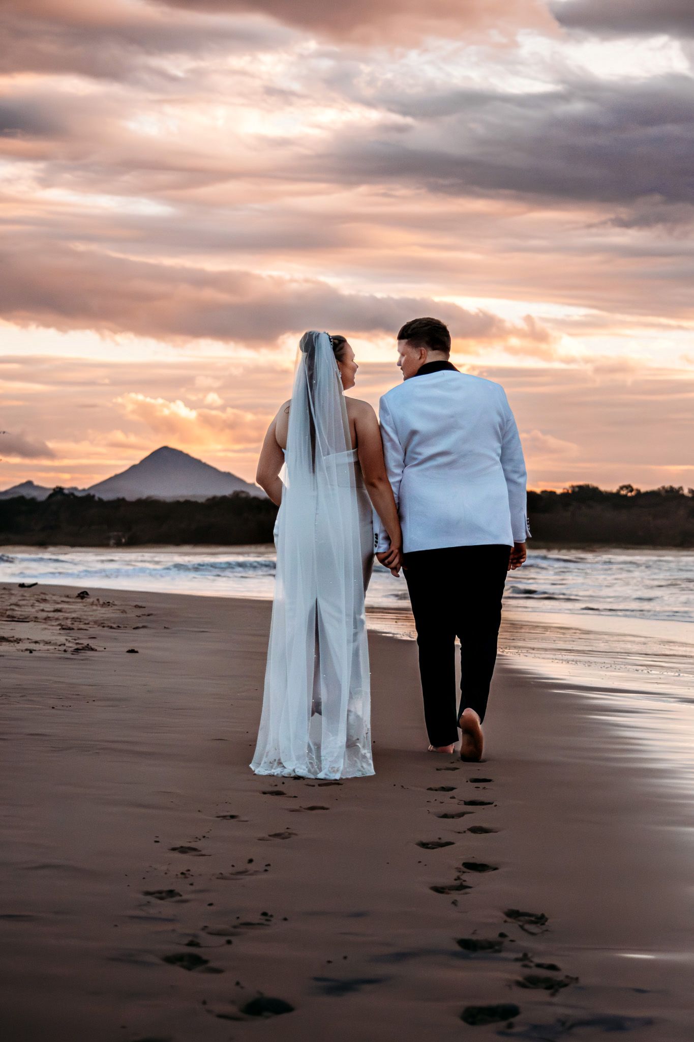 A couple walks along a beach at sunset leaving footprints in the sand.