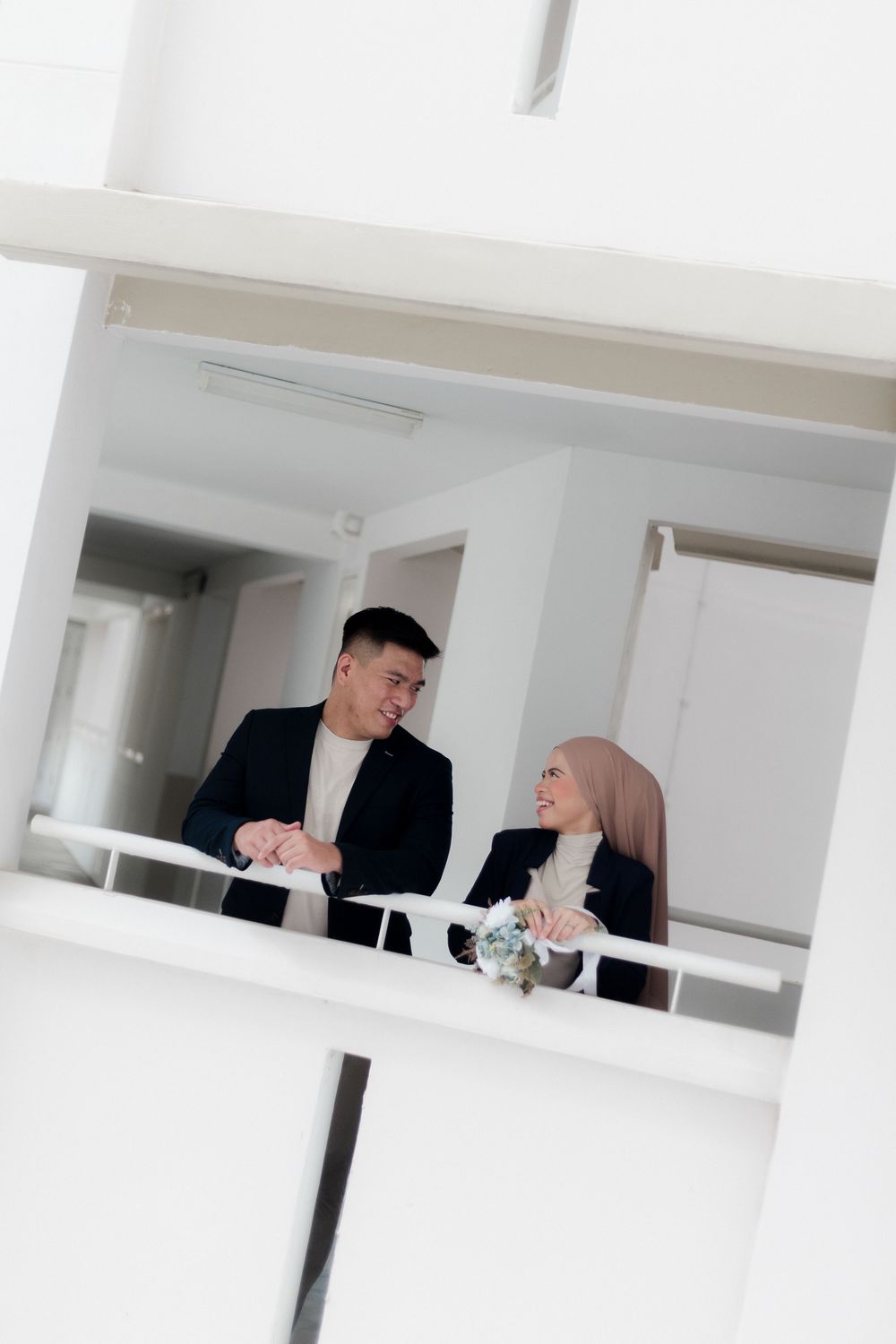 A smiling couple in matching black blazers stand together on a white balcony, looking at each other affectionately.