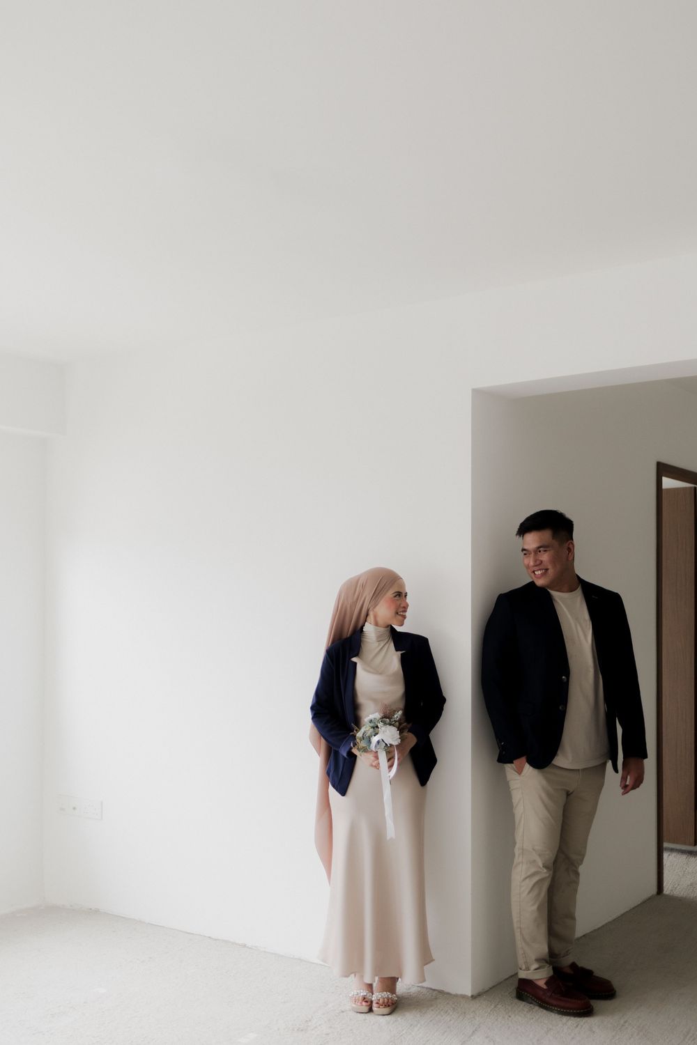 A couple in elegant attire poses in a minimalist white room, the woman holding a small bouquet while smiling at the man.