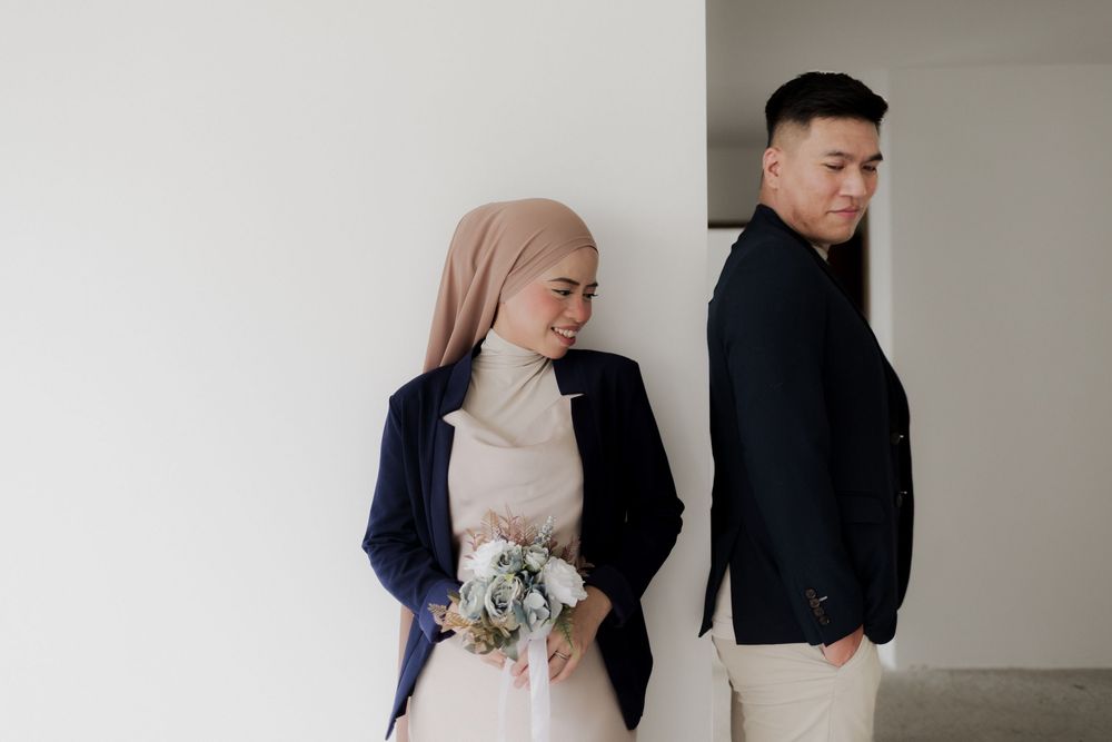 Muslim couple in matching navy blazers, bride holding white bouquet, posing back-to-back against white wall.