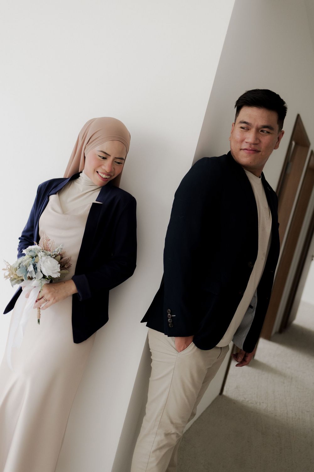 Muslim couple in matching navy blazers and beige outfits, woman in hijab holding bouquet, posing against white wall.