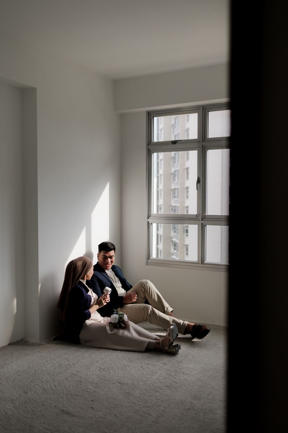 Couple sitting on floor of empty apartment near window, sharing a moment together in their new home.
