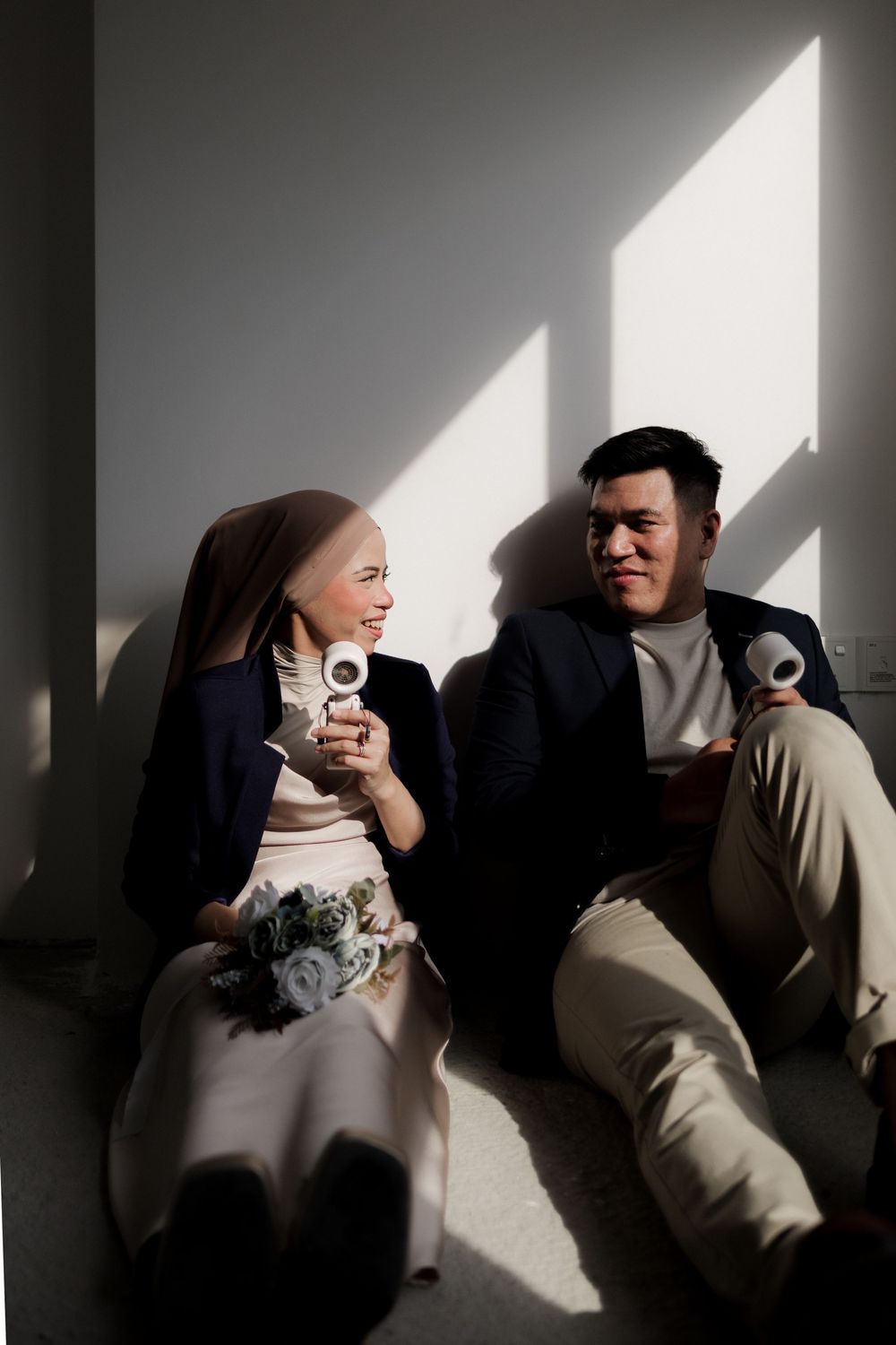 A couple sits on the floor in sunlight, smiling and holding coffee cups, dressed in elegant attire.